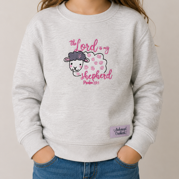 A child wearing a light heather gray sweatshirt with a pink and white graphic of a sheep and the text 'The Lord is my shepherd Psalm 23:1'. The child is also wearing blue jeans, standing against a neutral beige background.