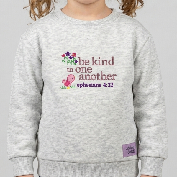 A young girl with curly brown hair wearing a light heather gray sweatshirt with a colorful graphic on the chest that reads 'be kind to one another ephesians 4:32'.