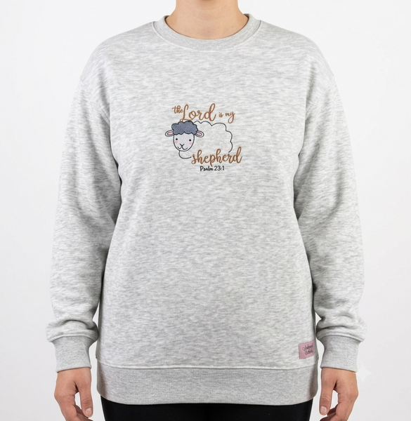 A person wearing a light heather gray crewneck sweatshirt with a graphic on the chest of a cartoon sheep wearing a blue cap and the text 'the lord my shepherd psalm 23:1'.