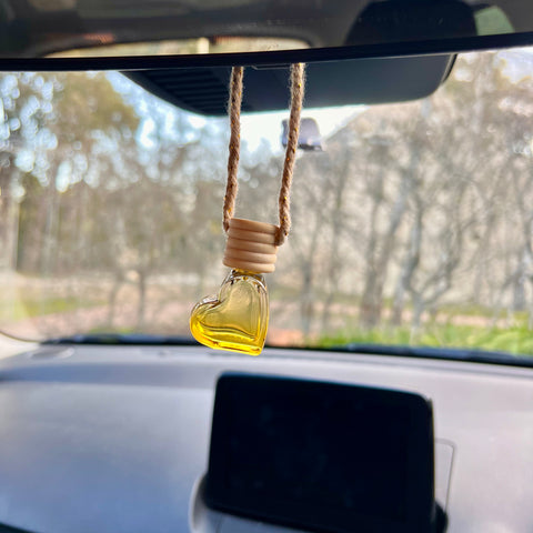 Heart-shaped car air freshener hanging from a car's rearview mirror with a blurred outdoor background.