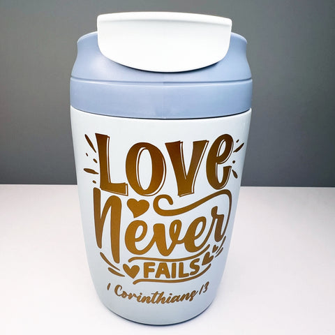White travel mug with blue lid featuring 'Love Never Fails' text on a gray background