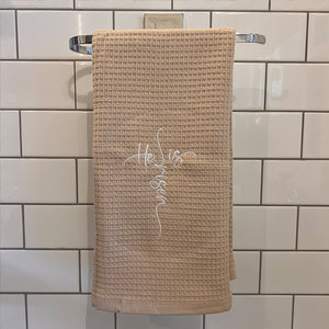 A beige kitchen towel with the phrase 'He is Risen' written in a red, cursive script, hanging on a metal rod against a tiled wall.